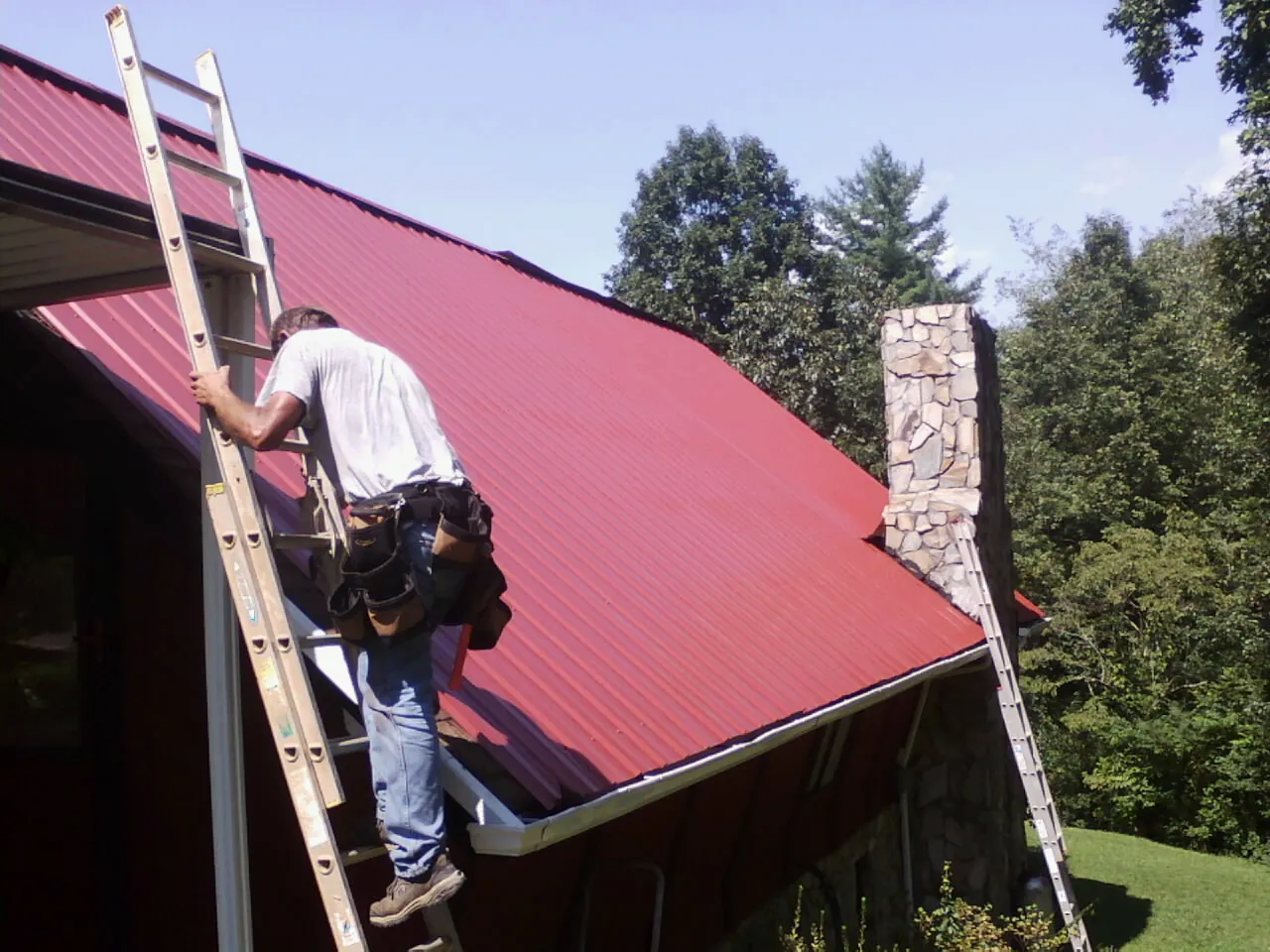 Roofer on a ladder beside a red metal roof and stone chimney — Soffit & Fascia Repair by Roofing Brothers in Asbury Park